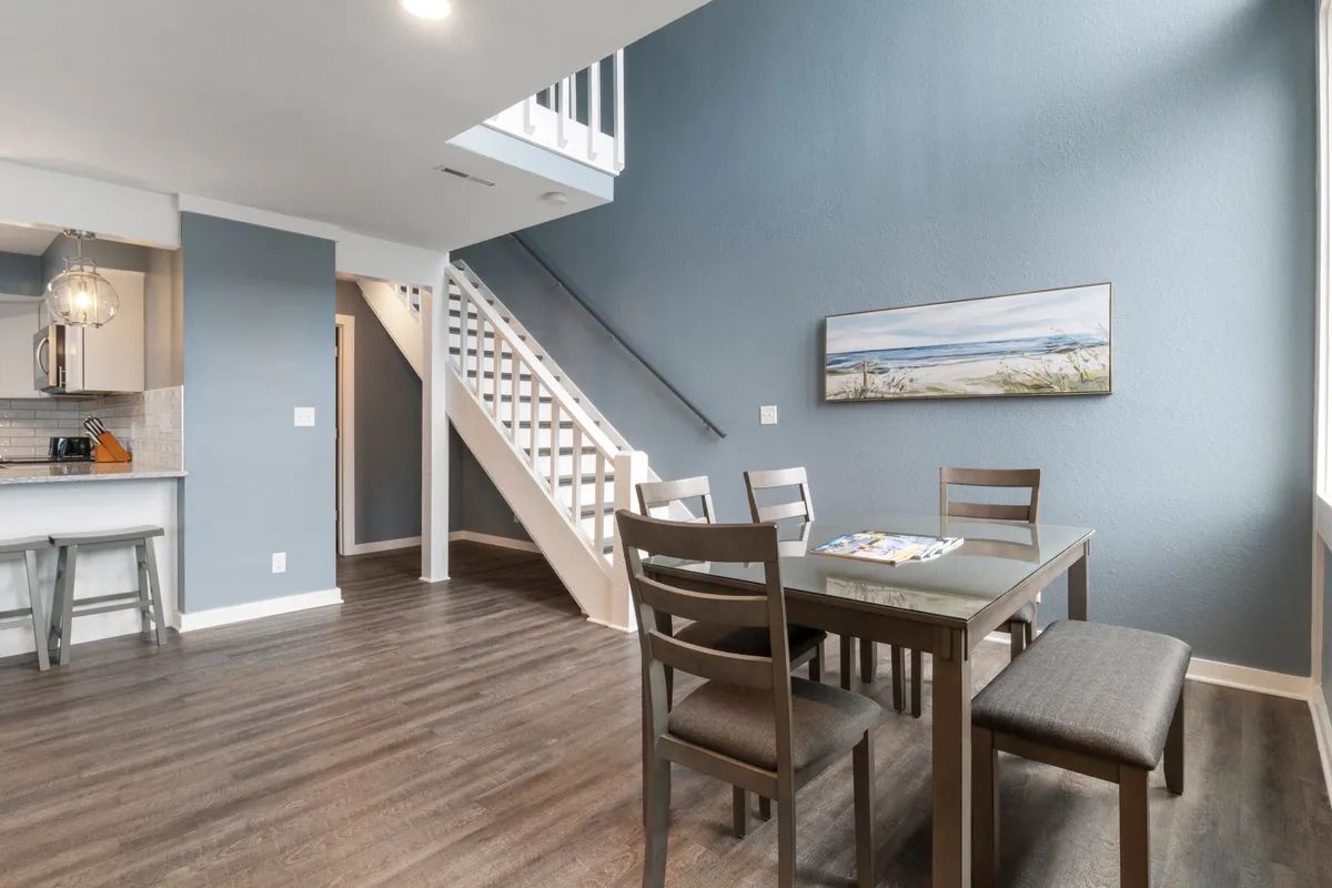 A cozy dining table adjacent to a staircase, with inviting wall art and a glimpse of the kitchen area. The scene suggests a welcoming space ideal for family meals, casual gatherings, or sharing morning coffee.
