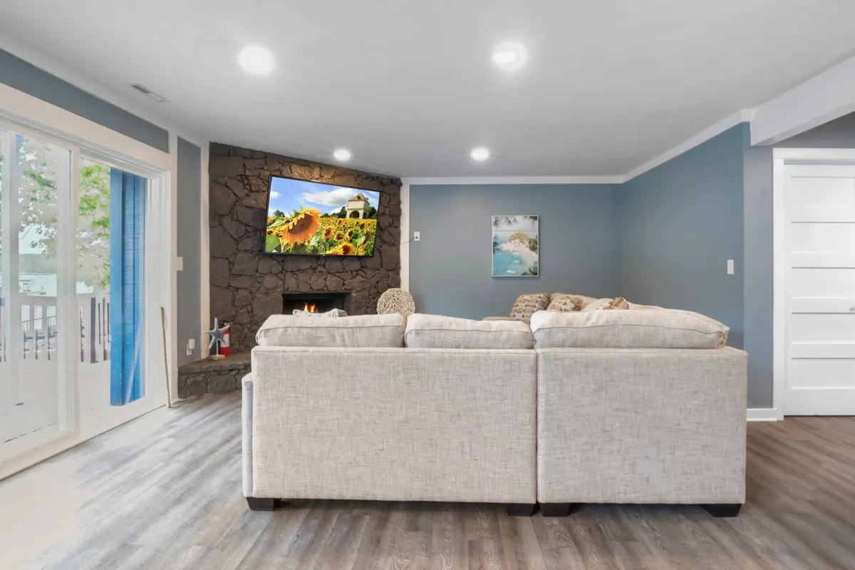 A bright and comfortable interior scene showing a large sectional sofa, a prominent fireplace, a television with a vibrant landscape image, and sliding glass doors leading outside. The scene exudes an inviting, lived-in feel, ideal for relaxing after pet walks or letting pets lounge close to family activities.