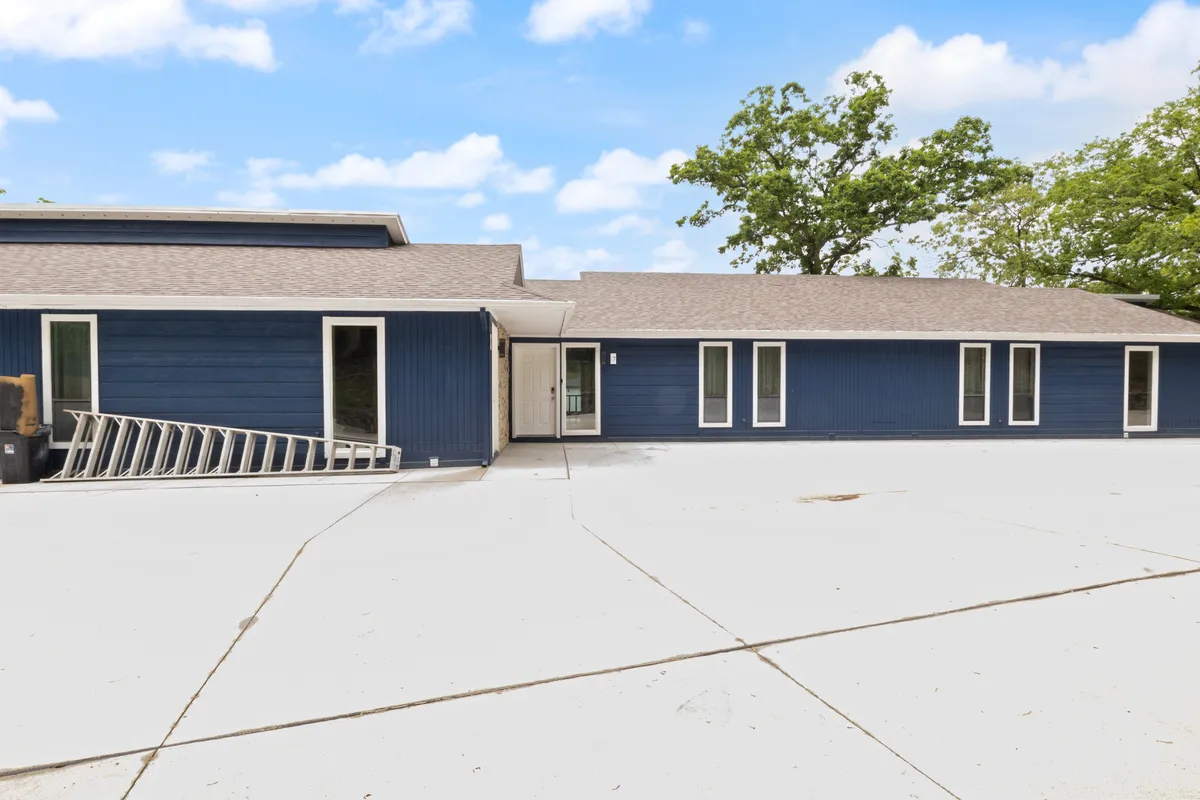 This image displays the blue exterior of the house, surrounded by a large driveway and greenery, with an inviting, spacious atmosphere ideal for outdoor family gatherings and lakeside activities. The scene suggests proximity to open air, relaxation, and group fun.