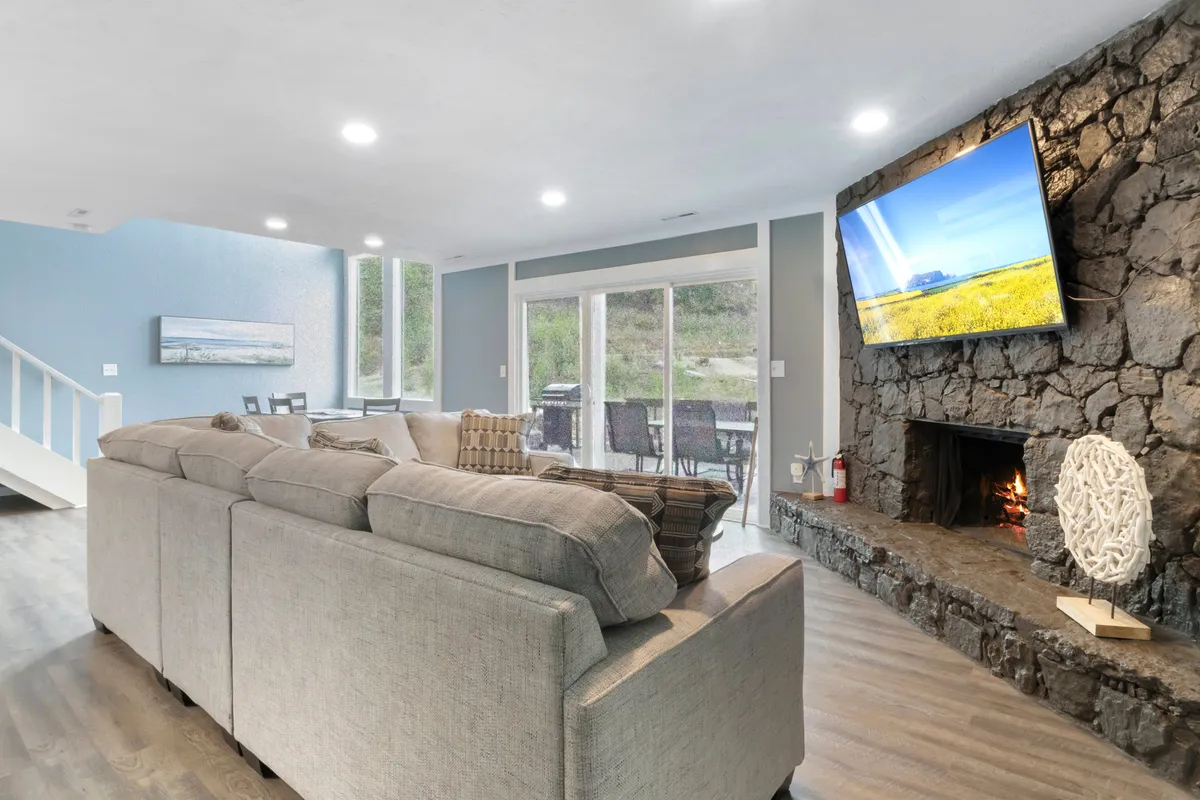 A warm, open living and dining space featuring a large sectional sofa, stone fireplace, flat-screen TV, and a dining area with a view. The room is filled with natural light, making it perfect for both relaxed meals and lively conversation.