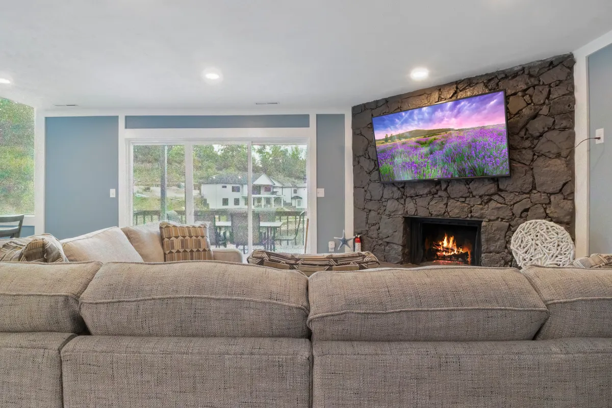 A cozy living room highlighted by a large sectional sofa, a fireplace, a television displaying scenic artwork, and windows with outdoor views. The abundance of soft seating and accessible windows suggests a pet-friendly haven where pets and guests can unwind with peace of mind.