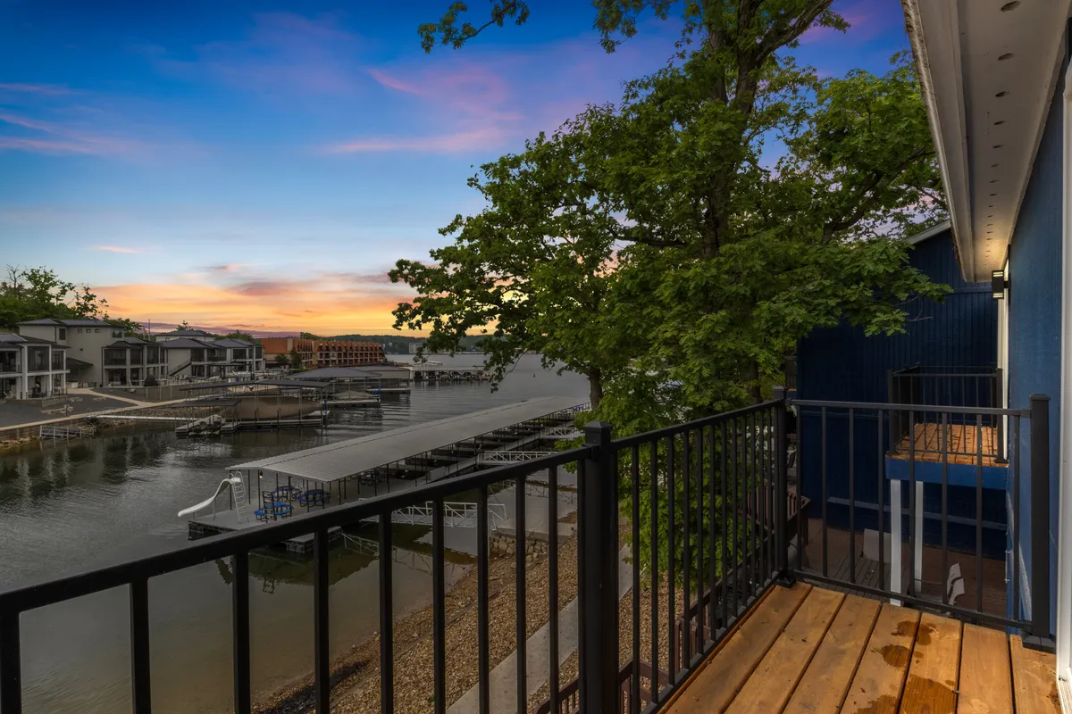 The image showcases a serene lakefront view from the condo's balcony, framed by lush trees and a glowing sunset sky reflected on calm water. The inviting scene suggests a tranquil setting ideal for relaxing, watching sunsets, and enjoying the peaceful cove atmosphere.