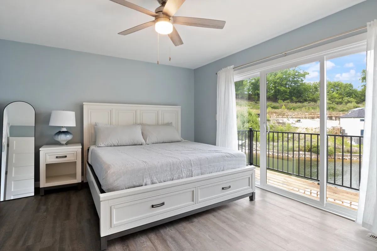 A serene bedroom featuring a plush bed adorned with soft linens, large windows allowing in natural light, a private balcony, and a tasteful mirror. The view from the windows suggests a tranquil connection with nature, making the space ideal for relaxing or enjoying a morning coffee.