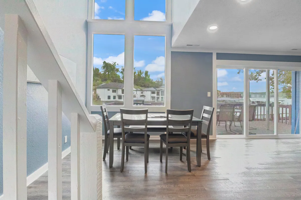 The image shows a bright dining area with a table set by large windows that offer a clear view of the outdoors, possibly of the lakeside or surrounding nature. Sunlight streams into the room, and the setting suggests a seamless connection between indoor comfort and the allure of the outdoors.